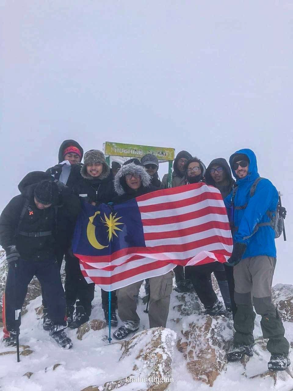 A visiting group holding a Malaysian flag at the Tidighin summit marker in snow